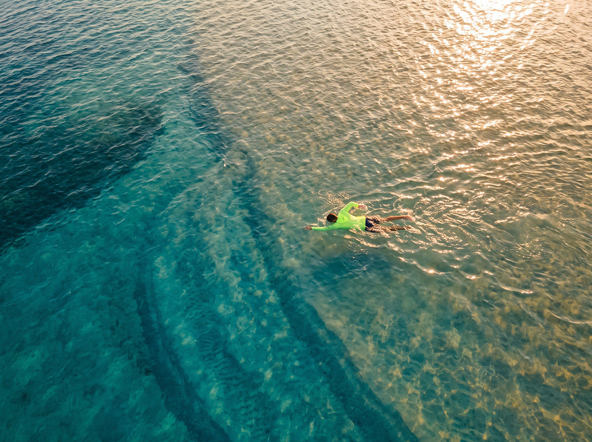 Aerial view of a swimmer in a bright green long-sleeve UPF sun shirt gliding through clear turquoise water at sunset, with sandbars and reef visible below the surface.