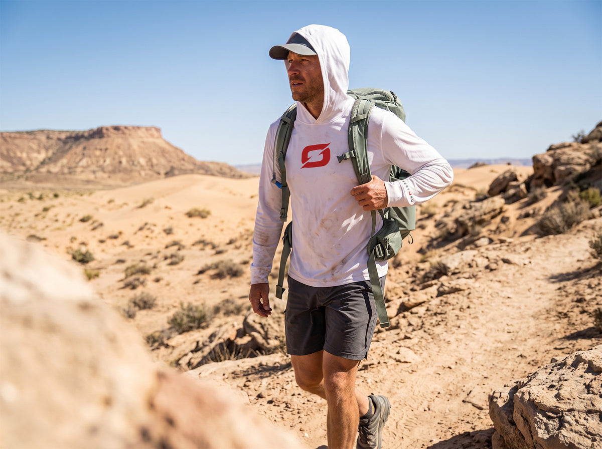 Hiker climbing a rocky desert trail under a clear blue sky, wearing a white Origin Gear Co. UPF 50+ hooded sun shirt with the orange logo on the chest, hood up under a cap and a green daypack.